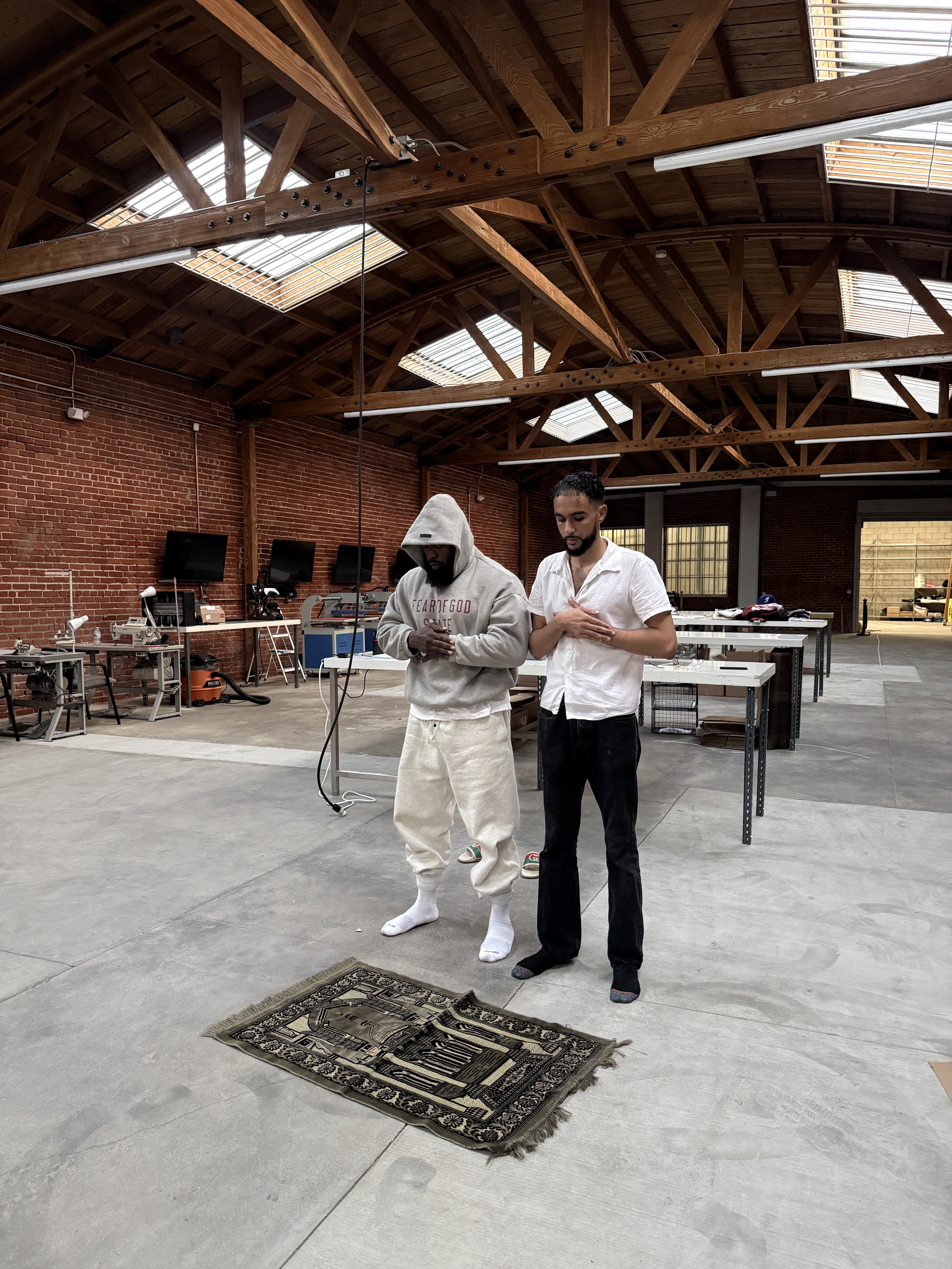 Two men standing in prayer beside a prayer rug in a brick-walled workspace
