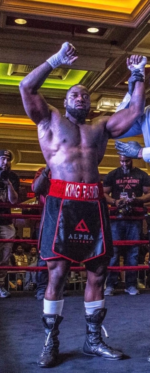Mike Rashid, wearing Alpha Shred boxing shorts, raising his arms in the ring